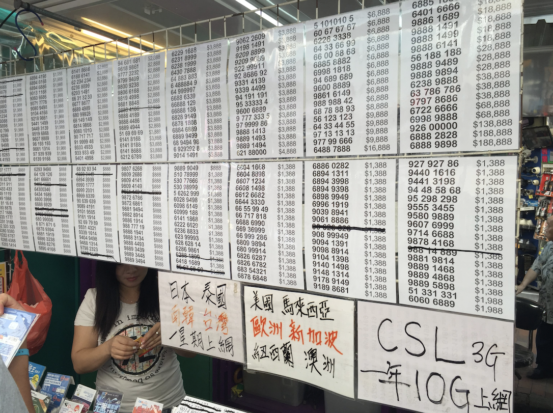 A wall covered in lists of phone numbers for sale with prices in Hong Kong dollars, with Chinese text below advertising different mobile carriers and services